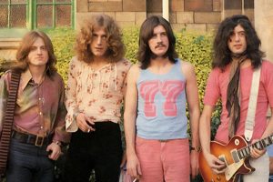 Four men stand outside in front of greenery and a brick building; one holds an electric guitar, and they wear casual 1970s clothing.