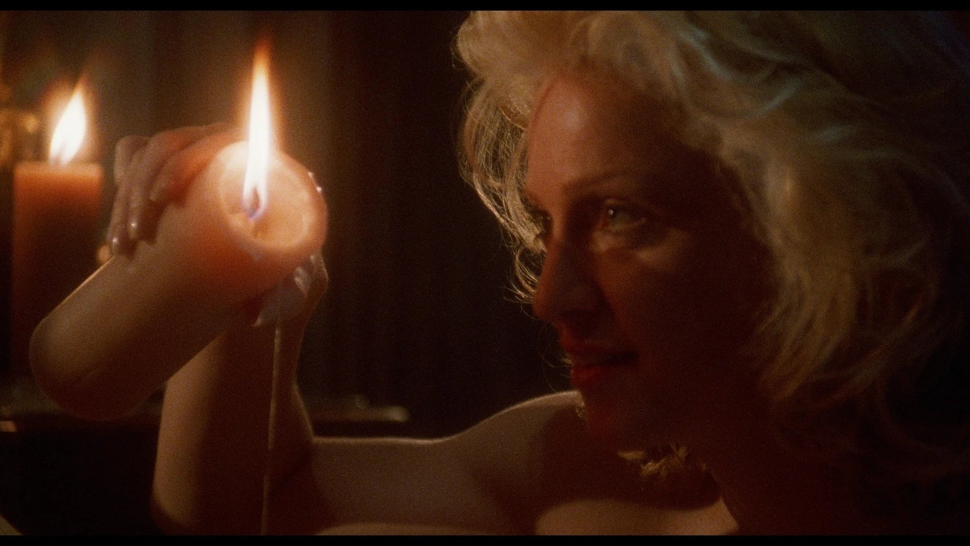 A person with blonde hair holds a lit candle, tilting it so melted wax drips down, with a dark background and another candle visible behind.