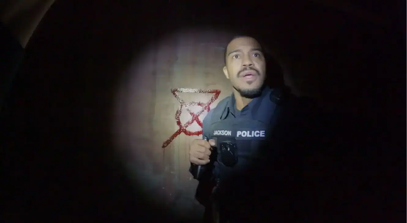 A police officer stands in the dark, illuminated by a flashlight, with red graffiti of an hourglass crossed out on the wall behind him.