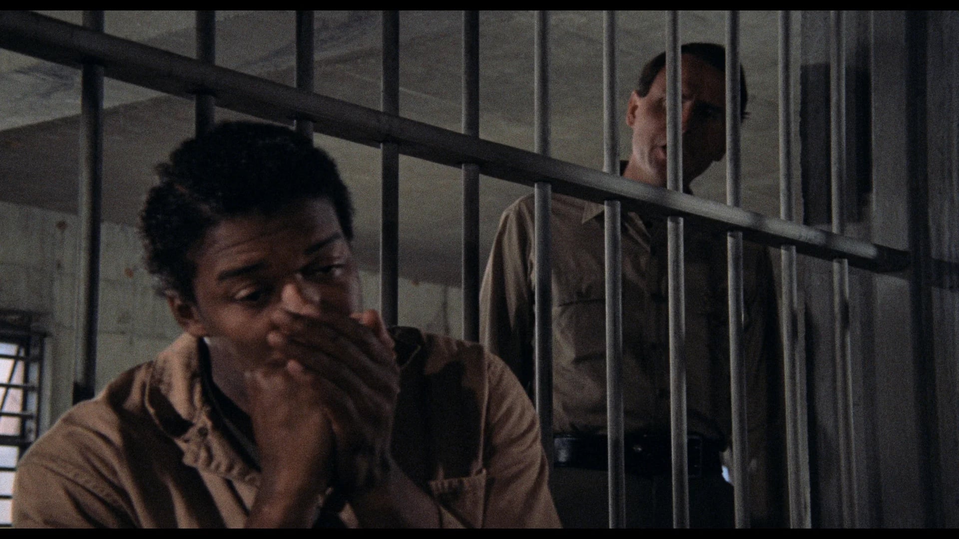 A man sits on a bench behind bars with his hands near his mouth, while a uniformed guard stands outside the cell looking at him.