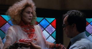 A woman with disheveled hair and a bloody, open neck wound stands in front of stained glass windows as a man in glasses holds her detached arm.