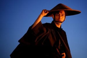 A person in traditional Japanese attire and a straw hat looks at the camera against a clear blue sky, holding the edge of the hat with one hand.