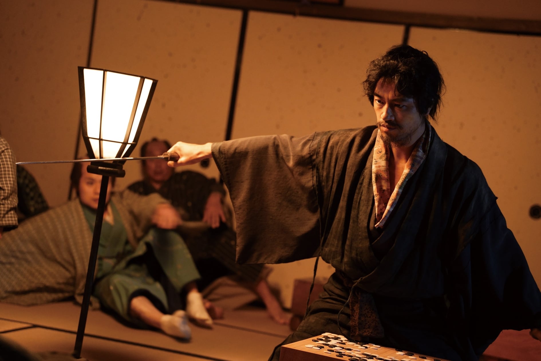 A man in traditional Japanese clothing points a sword at a lamp, with several people sitting on the floor in the background.