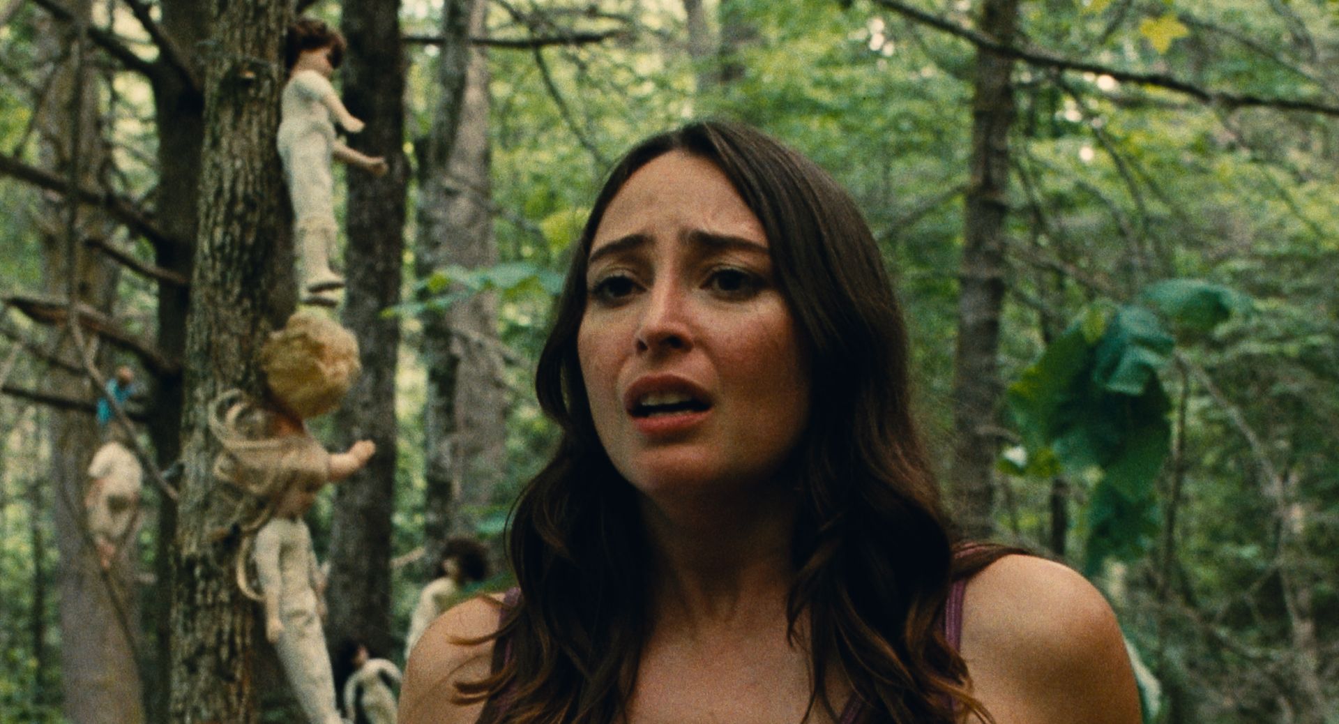 A woman with a worried expression stands in a forest; behind her, dolls are attached to a tree trunk.