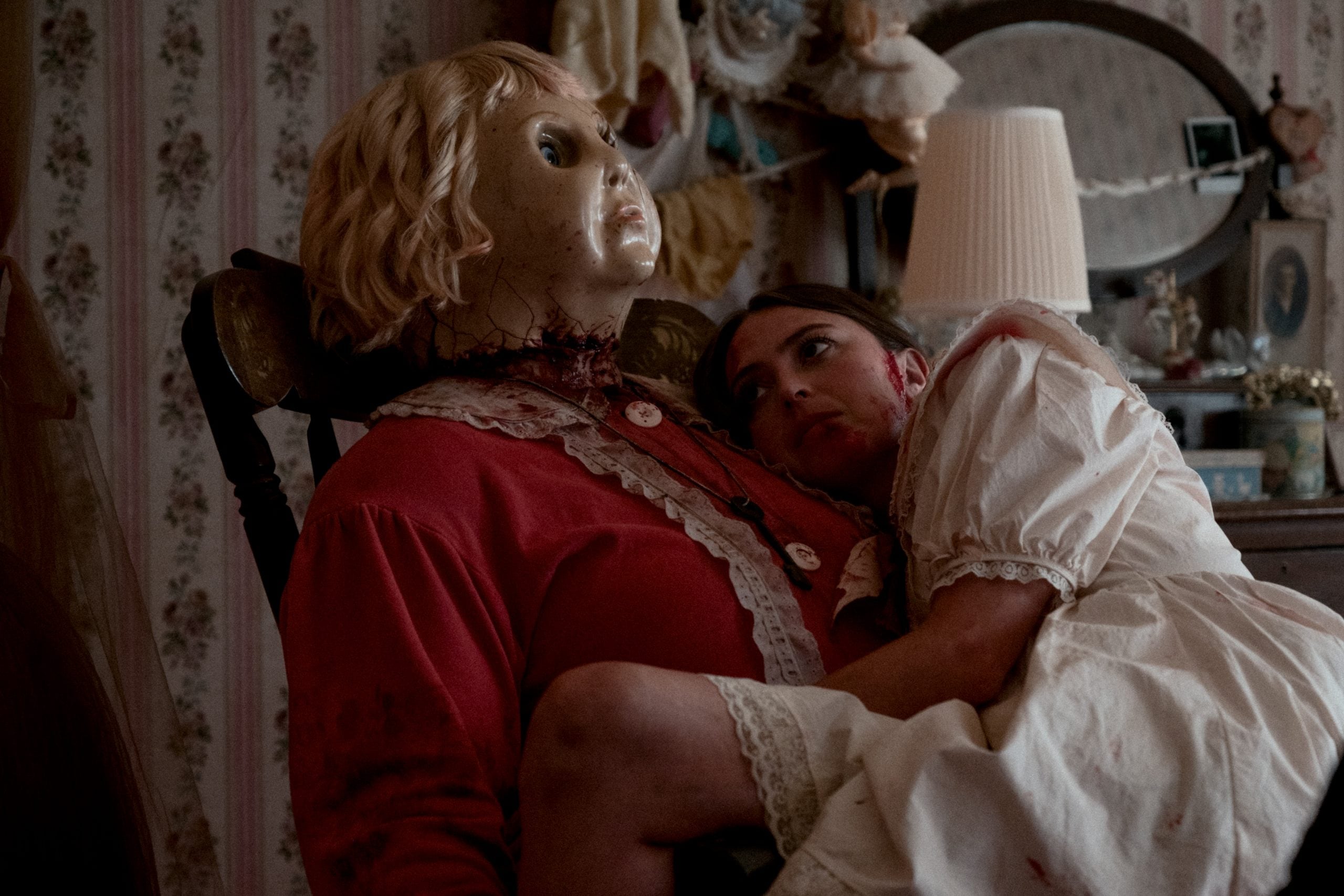 A woman in a white nightgown, with blood on her neck, leans against a large doll or mannequin with a cracked face and a severed head, in a dimly lit, old-fashioned bedroom.