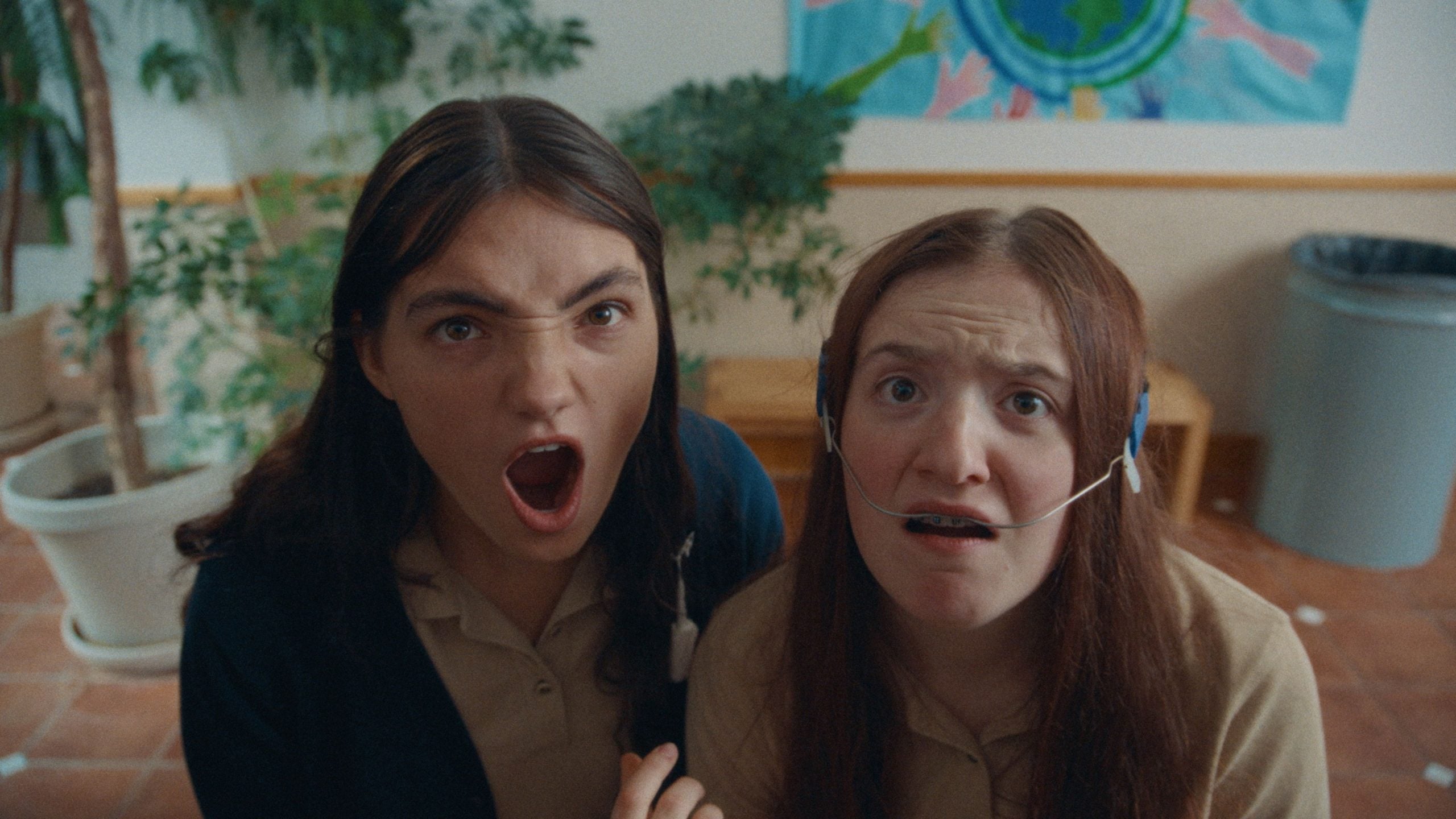 Two young women in tan shirts look up at the camera indoors; one appears shocked with mouth open, the other has a nasal cannula and a concerned expression.