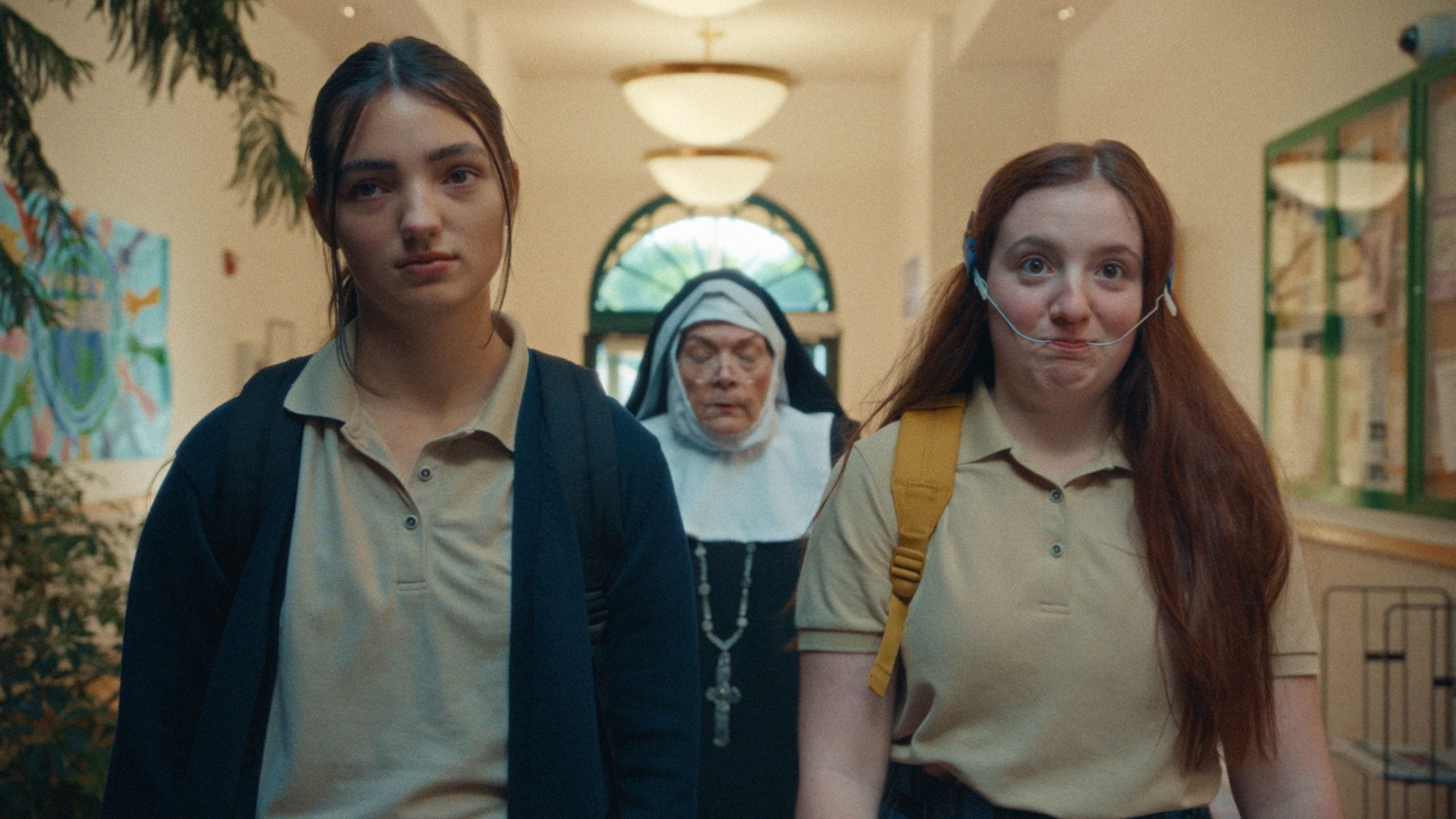 Two teenage girls in school uniforms walk down a hallway, one wearing a headset and making a face, with a nun in a habit following behind them.