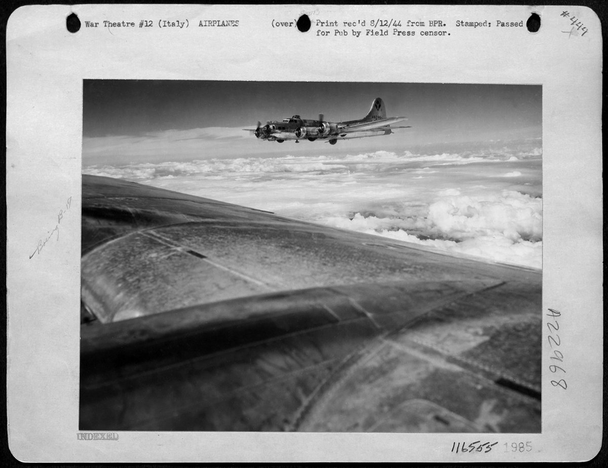 A B-17 bomber in flight above the clouds, photographed from another aircraft during World War II; black and white image with archival notes on the border.