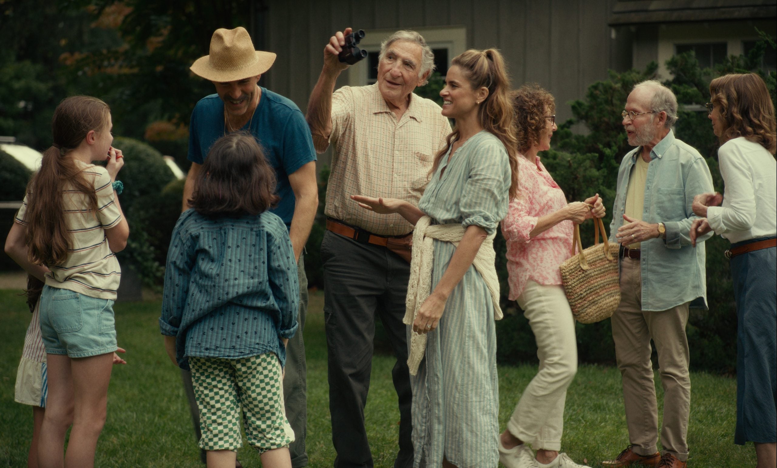 A group of people of various ages stand together outdoors on grass, smiling and talking while one person takes a photo with a camera.