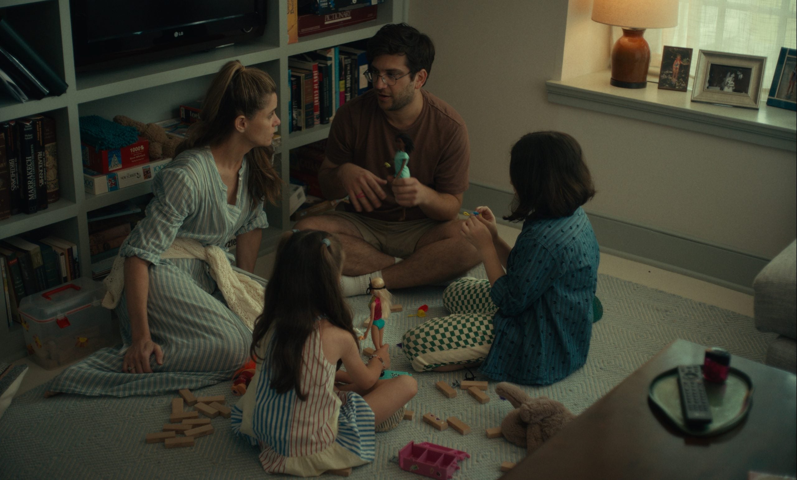 A family of four sits on the floor in a living room, playing with toys and wooden blocks near a bookshelf and a window.