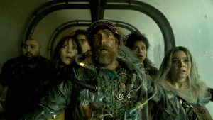 Five people, wearing distressed and improvised outfits, stand closely together in a dimly lit tunnel, looking ahead with expressions of shock and concern.