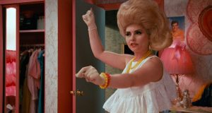 A woman with a large beehive hairstyle and vintage clothing in a colorful, retro bedroom.