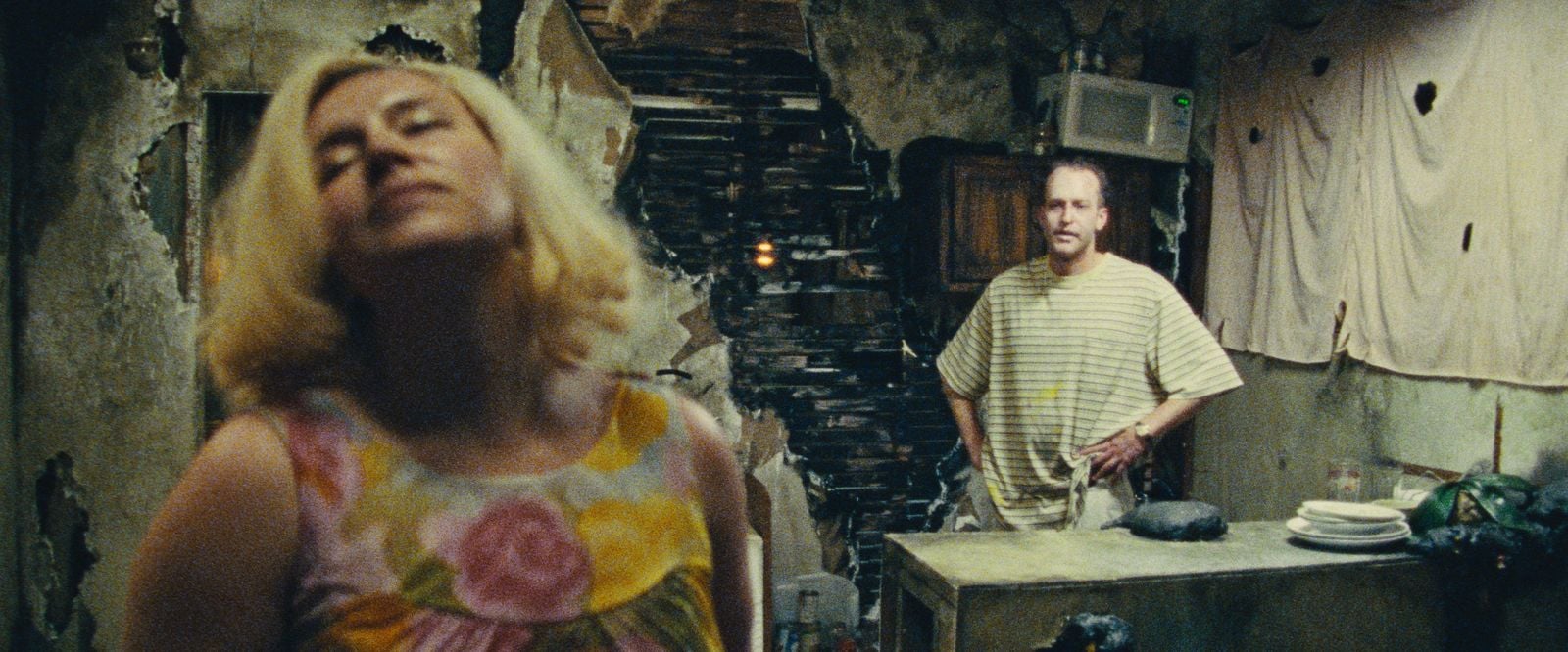 A woman in a floral dress stands with her head tilted in the foreground, while a man in a striped shirt stands in a rundown kitchen with peeling walls and exposed bricks.