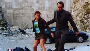 A man in a suit and a young woman in casual clothes walk through a rocky area with debris, with a black car parked behind them.
