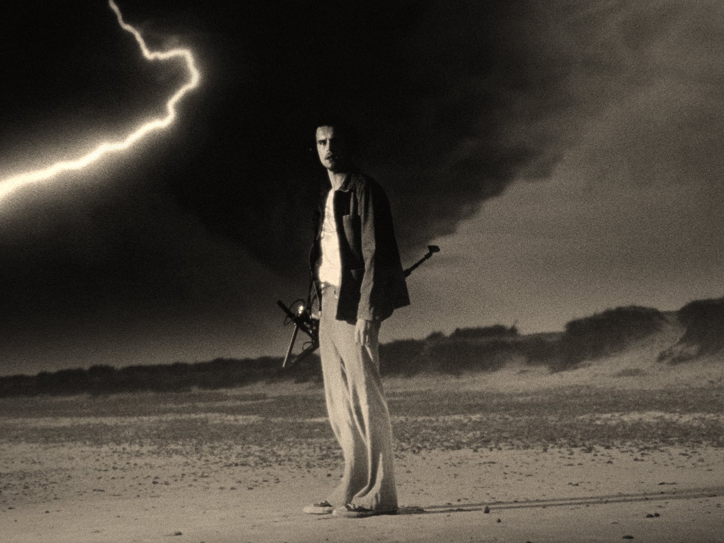 A person stands on a beach holding a sword, with a dark stormy sky and a lightning bolt in the background.