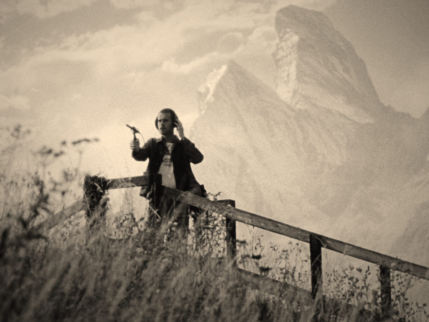 Person standing on a wooden fence in a grassy area, aiming a slingshot, with large, rugged mountains in the background under a cloudy sky.