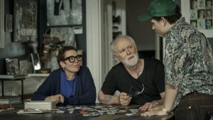 Three people are gathered around a table covered with buttons, having a conversation in a room filled with artwork and photographs.