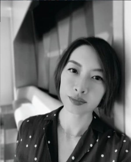 A woman with short hair and a polka dot shirt stands indoors, leaning against a wall, looking at the camera in a black and white photo.