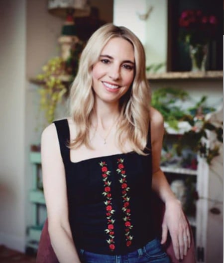A woman with long blonde hair, wearing a black sleeveless top with floral embroidery, sits on a chair in a room decorated with plants and shelves.
