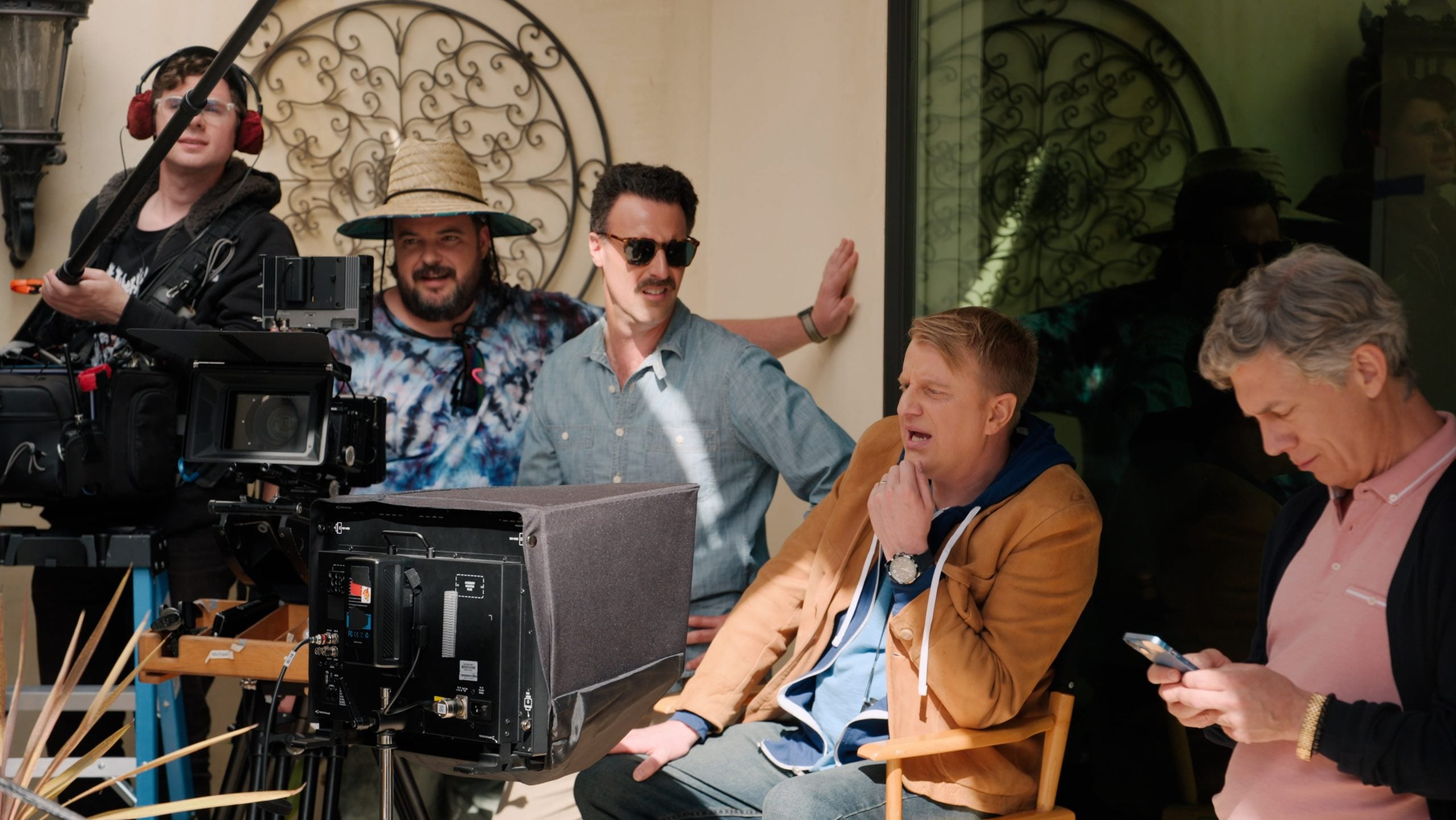Four men sit and stand around film equipment outdoors, one operating a camera, another checking a phone, while others look toward the set. Decorative ironwork is visible in the background.