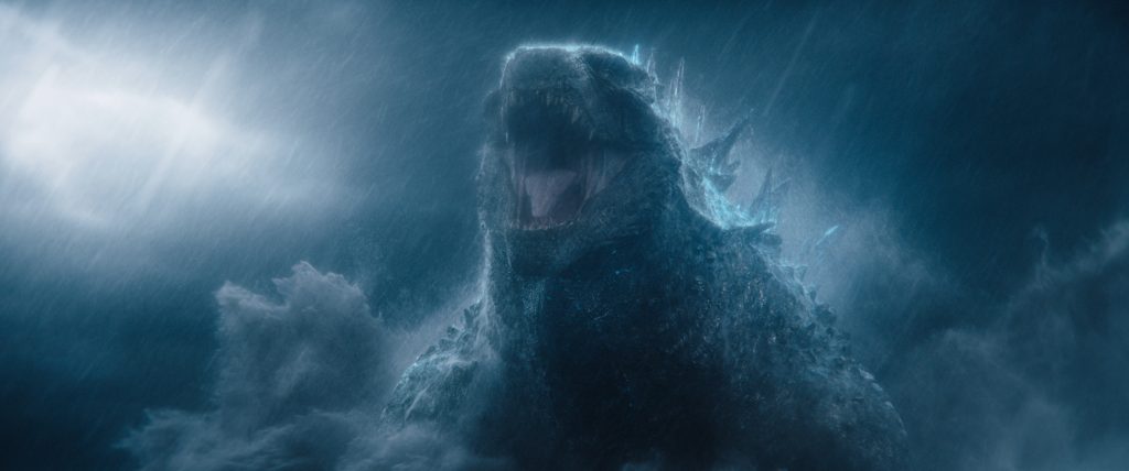 A large, roaring Godzilla stands amid rain and mist, illuminated by lightning in a dark, stormy atmosphere.