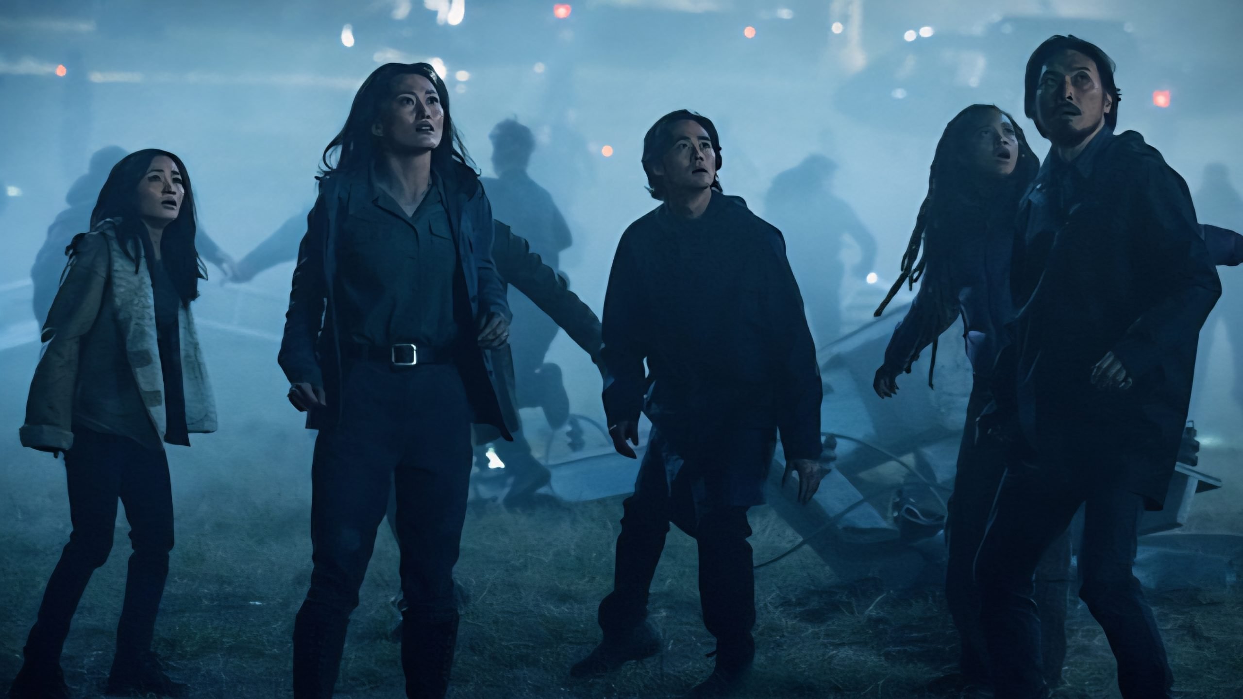 Five people stand outdoors at night, looking up with concerned expressions as debris and fog surround them, illuminated by blue-tinted lights.