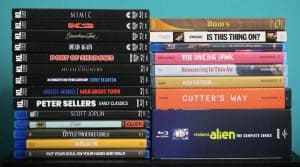 A stack of black DVD cases on the left and colorful DVDs and Blu-rays on the right, all with visible spines showing various movie and TV titles.