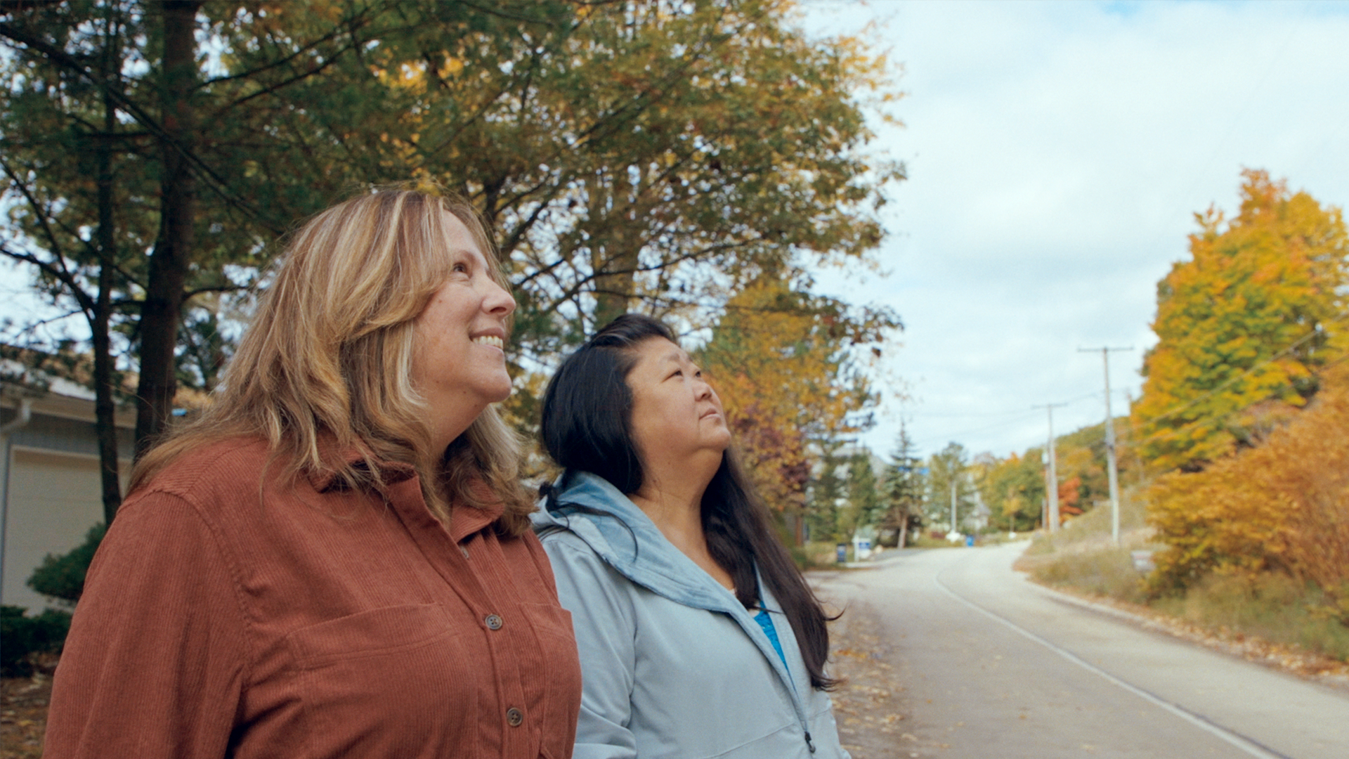 Two women stand by a road lined with autumn trees, looking up and smiling as they enjoy the outdoor scenery on a cloudy day.