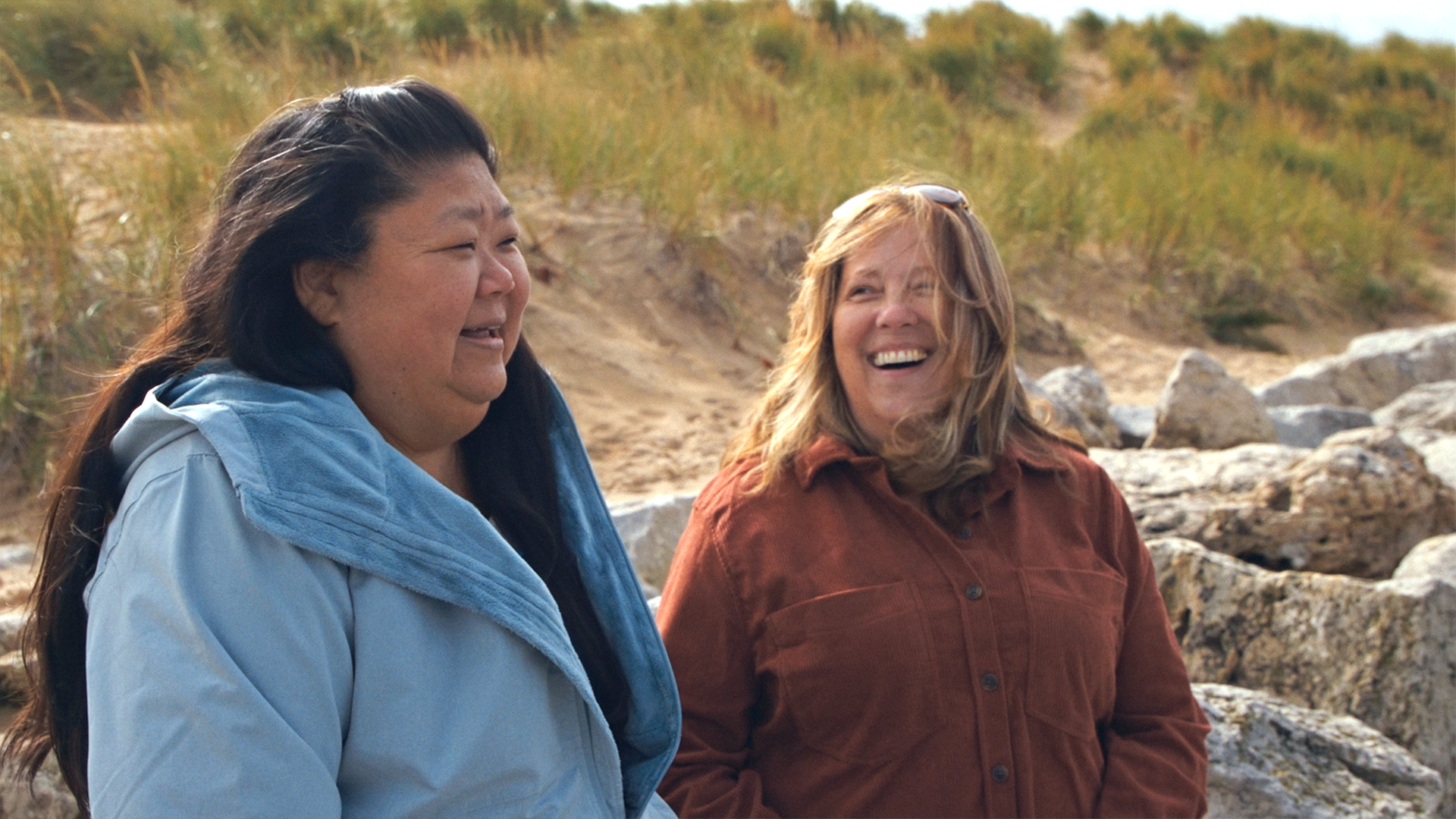 Two women stand outdoors, smiling and laughing together near rocks and grassy sand dunes on a cloudy day.