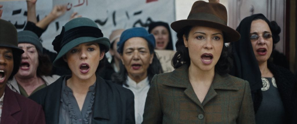 A group of women wearing hats and period clothing stand together, appearing to protest or shout, with a banner visible in the background.