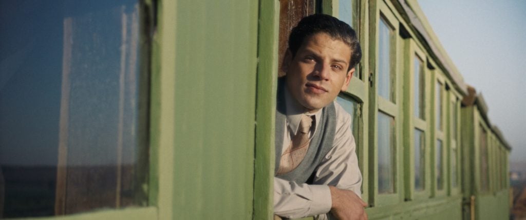 A man in a suit leans out of the window of a green train car, looking into the distance on a clear day.