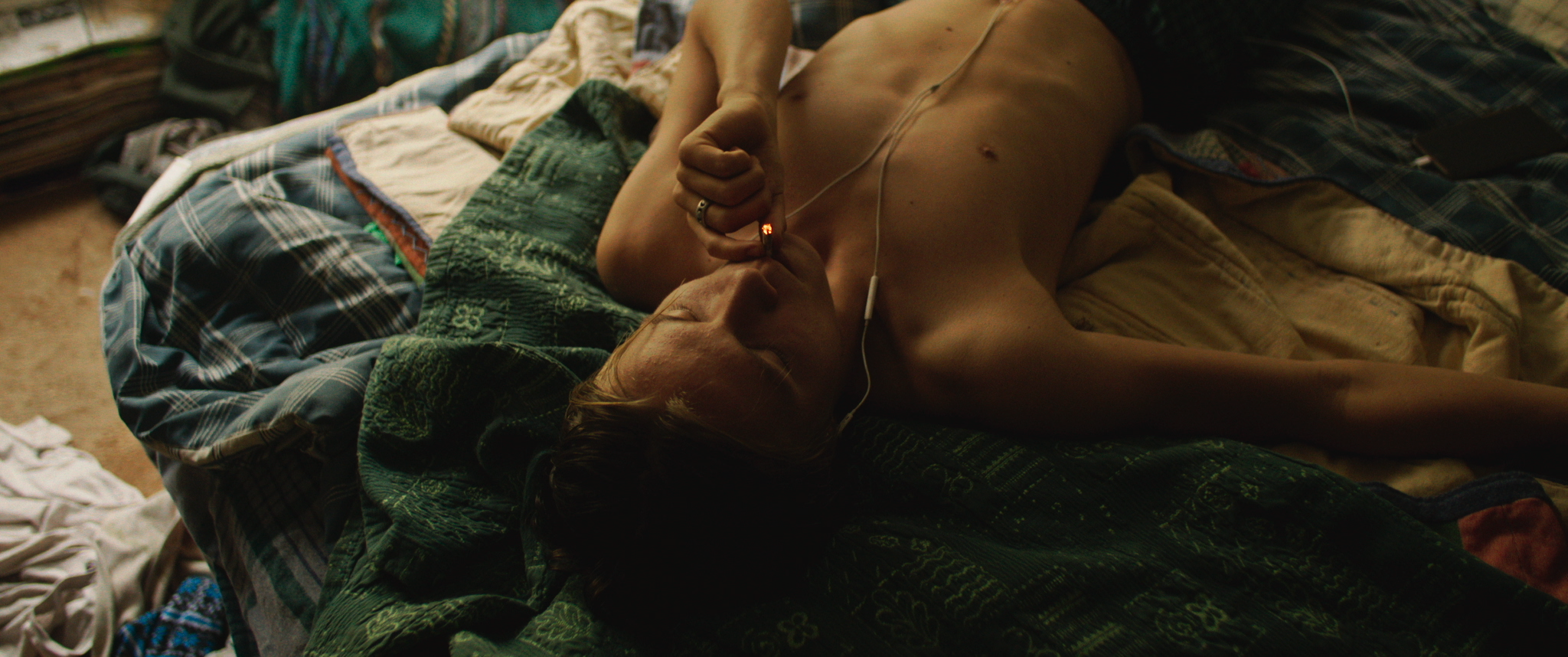 A shirtless person lies on a messy bed, listening to earphones and smoking, with a dimly lit room and clothes scattered around.