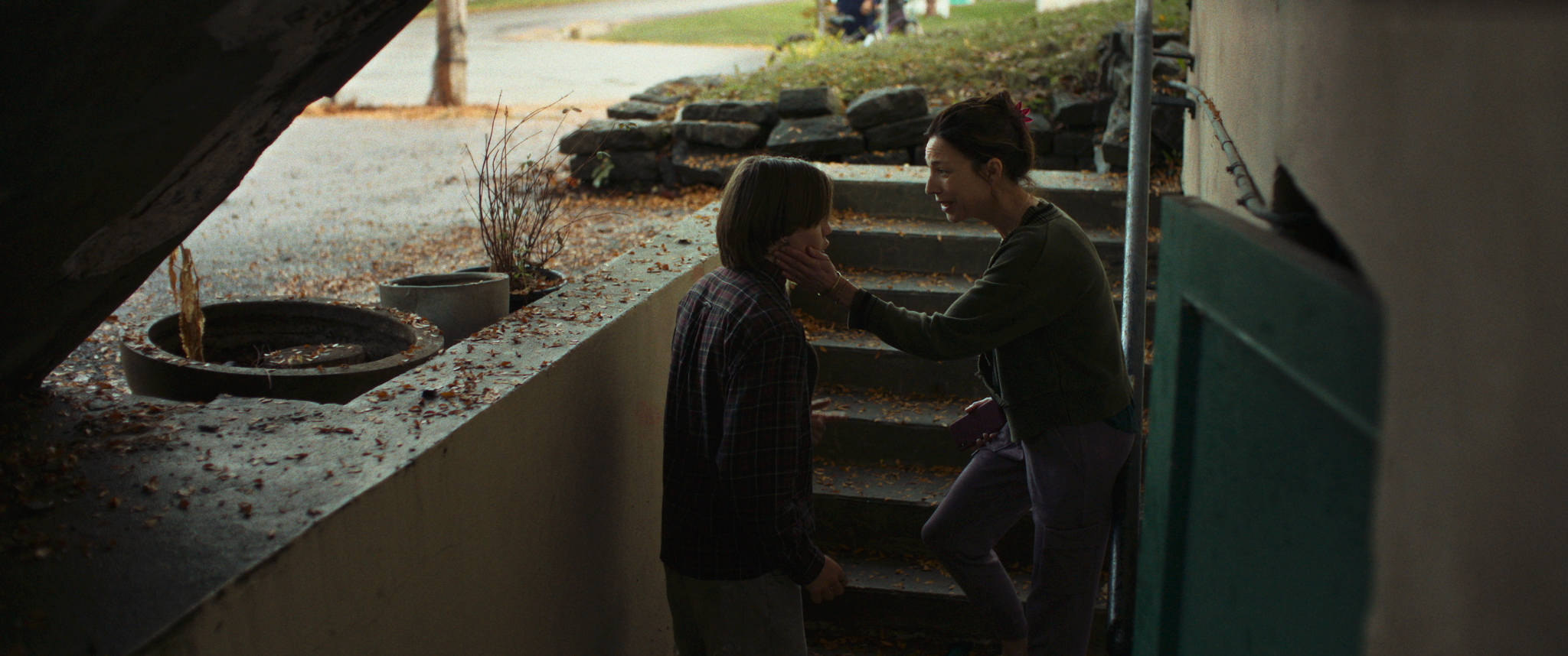 A woman stands on outdoor steps, holding a young person's face with both hands, in a shaded, leaf-strewn area near a green door.