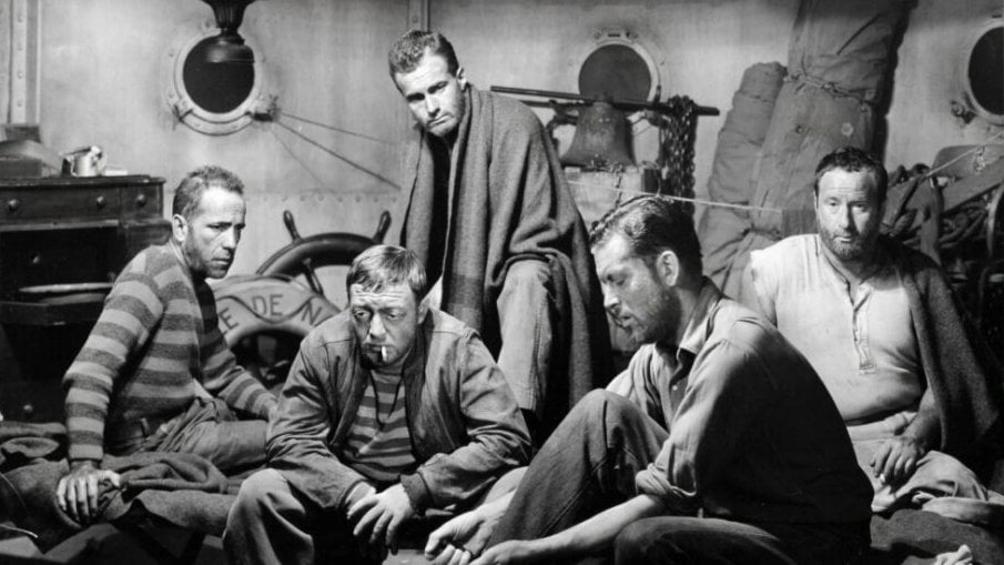 Five men sit or crouch in a dimly lit, cluttered ship cabin, wearing expressions of concern or exhaustion; portholes and various ship equipment are visible in the background.