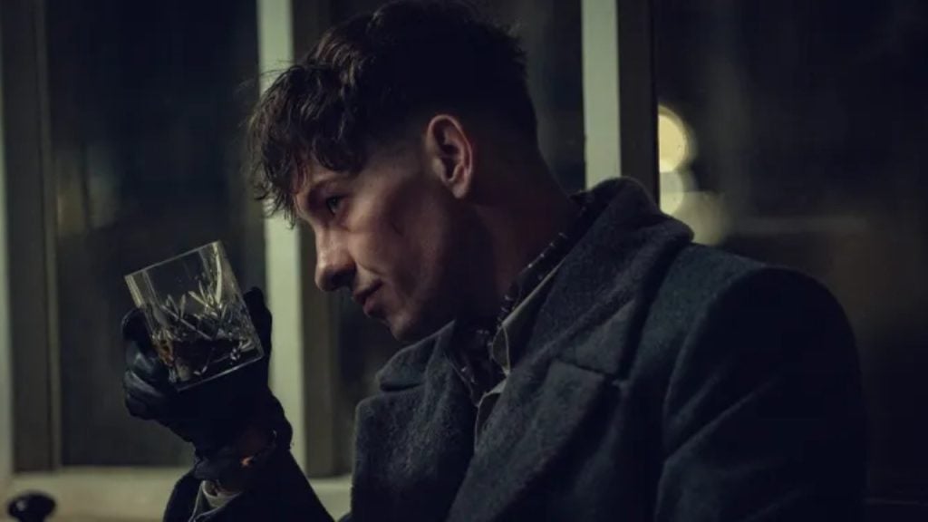 A man wearing a coat and gloves sits indoors at night, holding up a glass of dark liquid and looking at it thoughtfully.