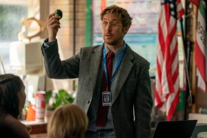 A man in a suit and lanyard holds up a small globe while speaking to a group in a classroom with American and California flags in the background.