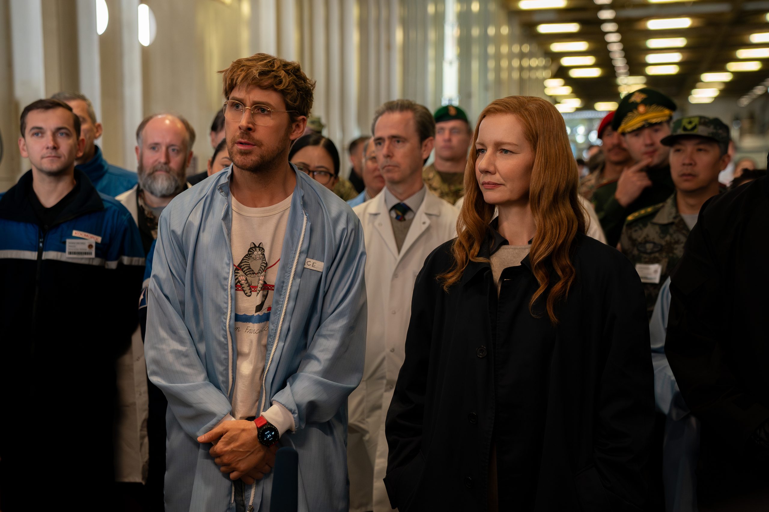 A group of people, including scientists in lab coats and others in casual or military attire, stand together indoors, listening attentively.