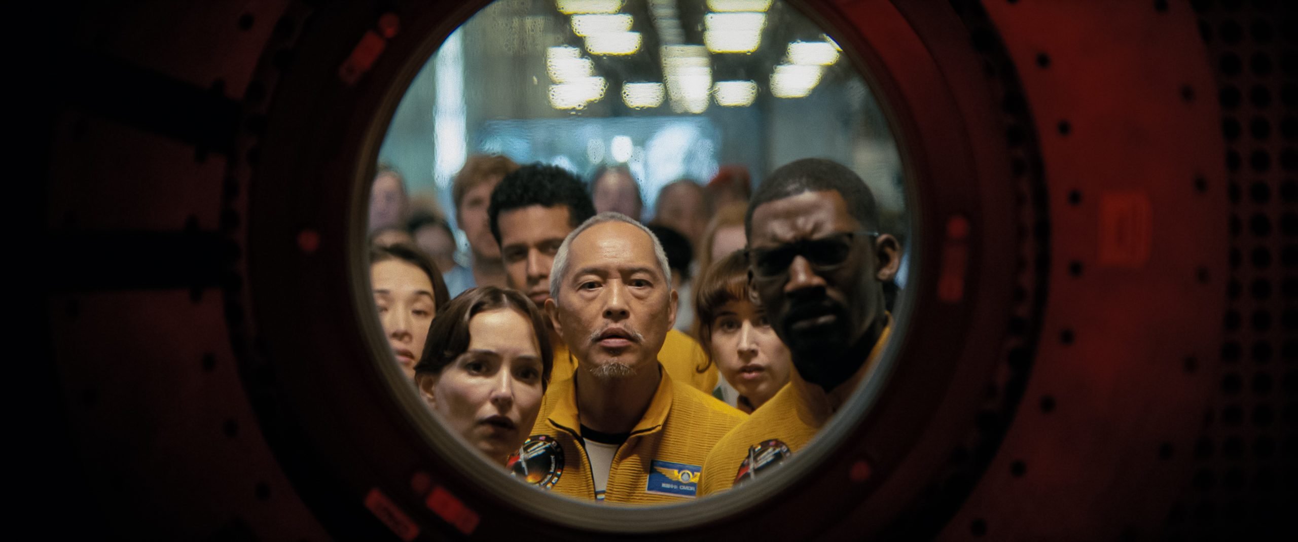 A group of people in yellow uniforms closely peer through a circular window or hatch, appearing attentive and serious.