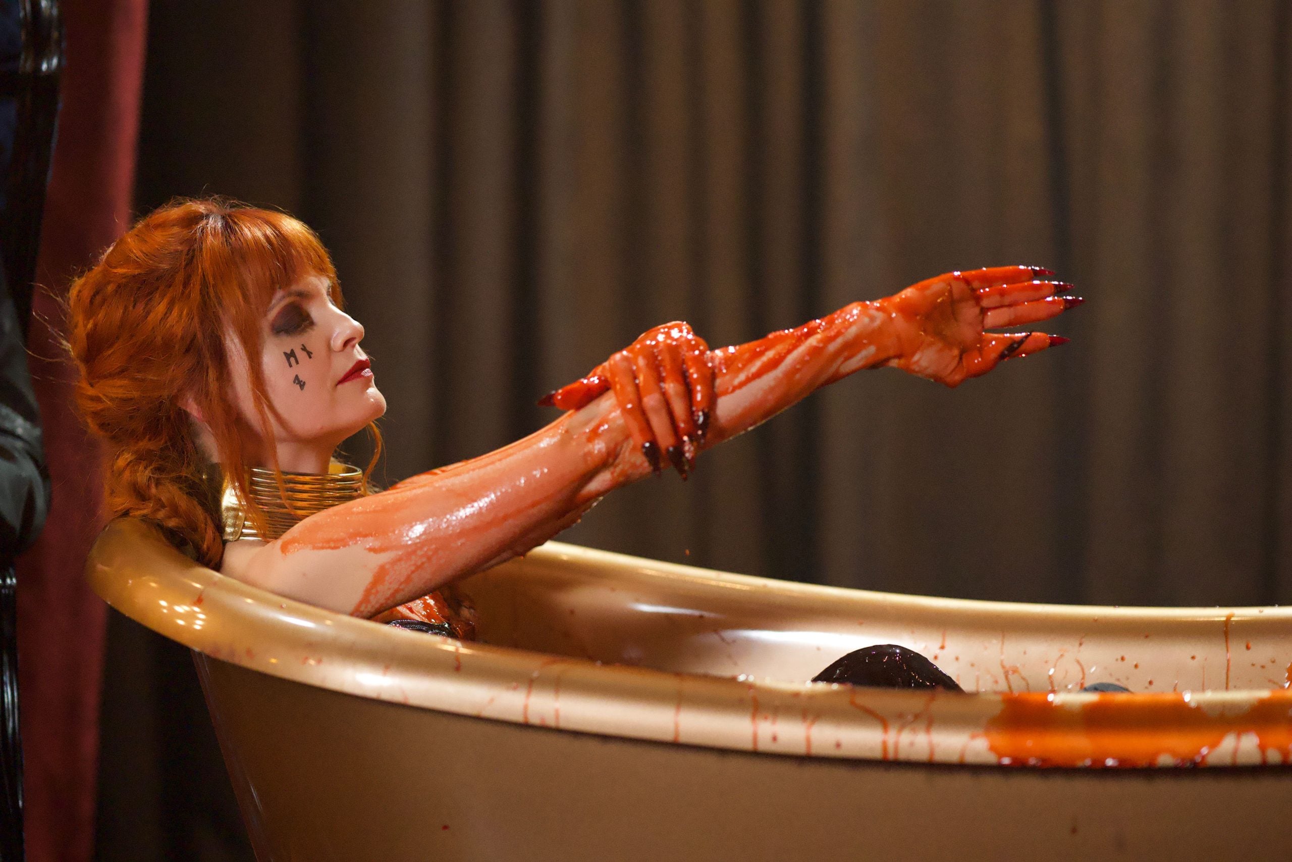 A person with red hair and black markings on their face sits in a gold bathtub, covered in red liquid, with arms extended and long black nails.