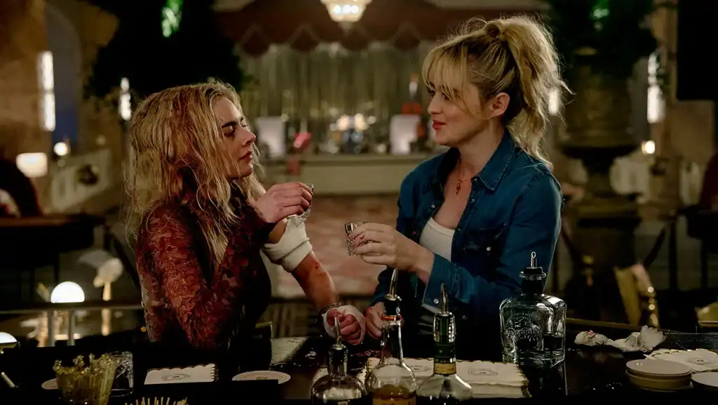 Two women sit at a bar facing each other, holding up shot glasses as if toasting, with bottles and bar tools on the counter in front of them.