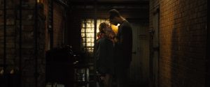 A man and woman stand closely facing each other in a dimly lit brick hallway, with the man gently holding the woman's face and leaning toward her.