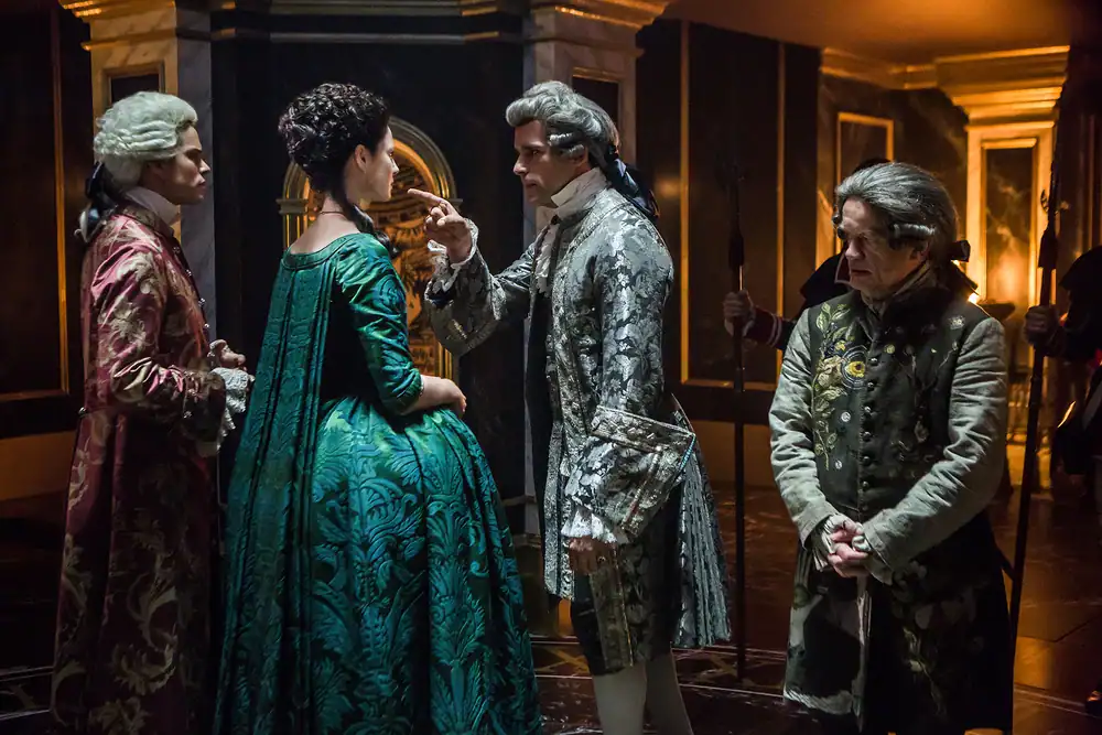 Four people in elaborate 18th-century costumes stand in a richly decorated room; one man gestures towards a woman in a green gown while two other men observe.