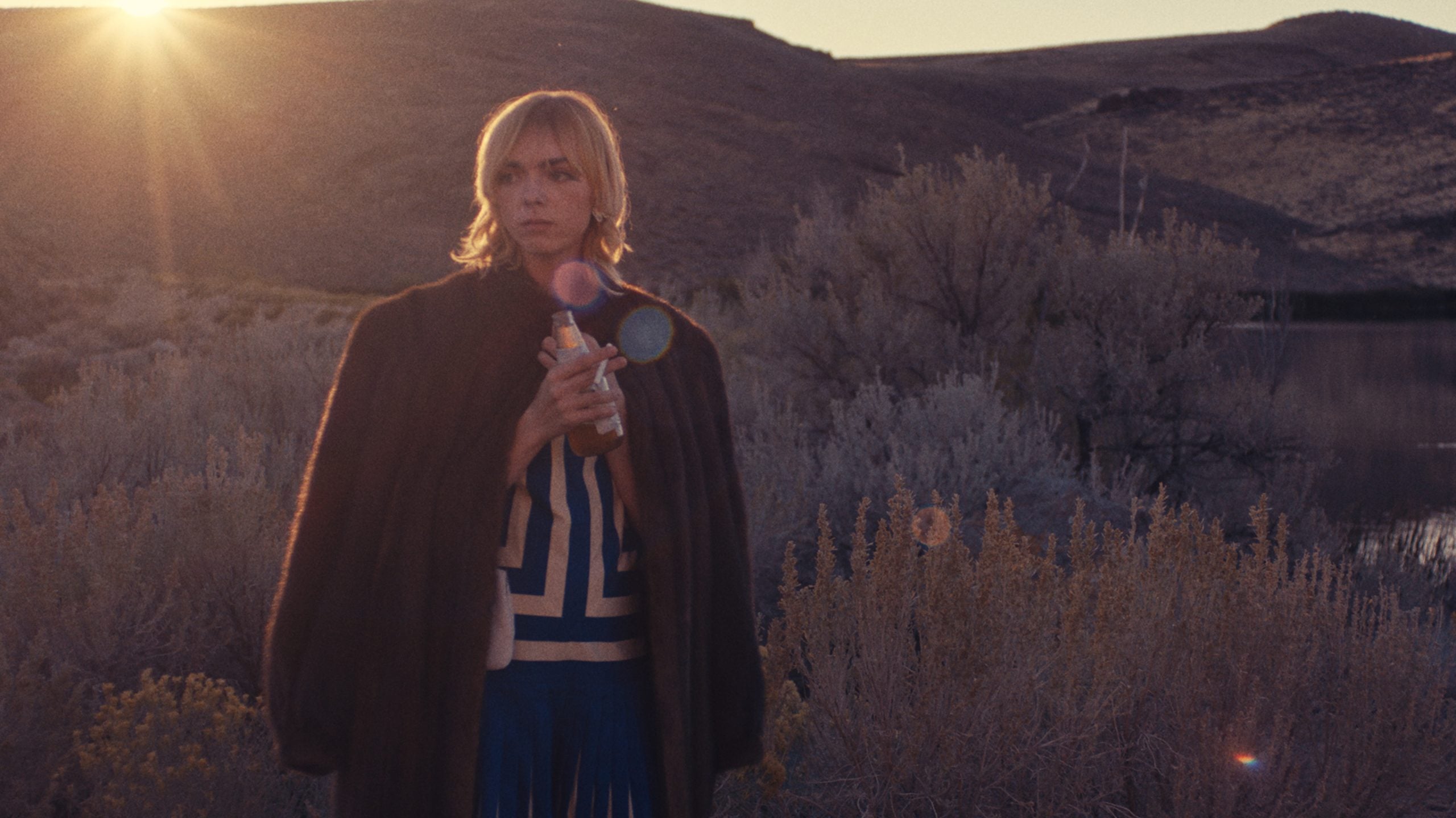 A person wearing a patterned shirt and dark coat stands in a dry, shrubby landscape at sunset, holding a drink with hills and a body of water in the background.