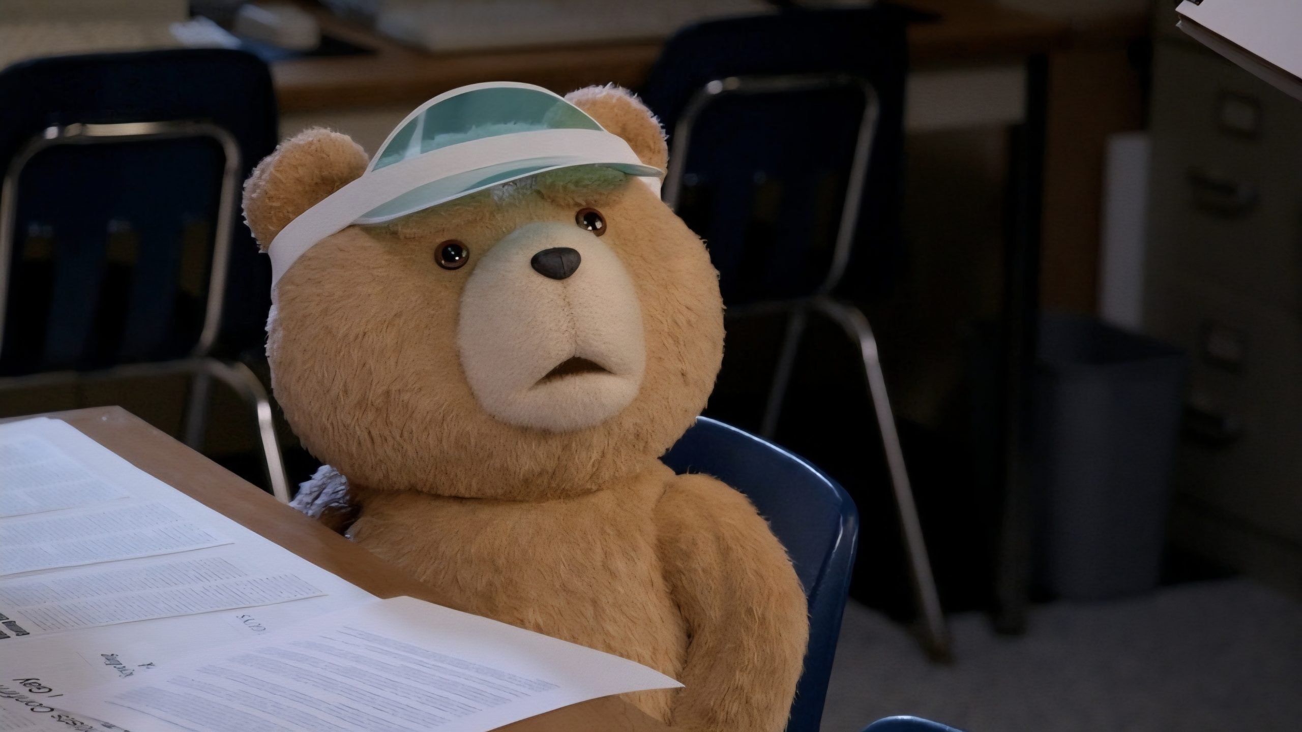 A teddy bear wearing a visor sits at a desk with papers, in a classroom or office setting.