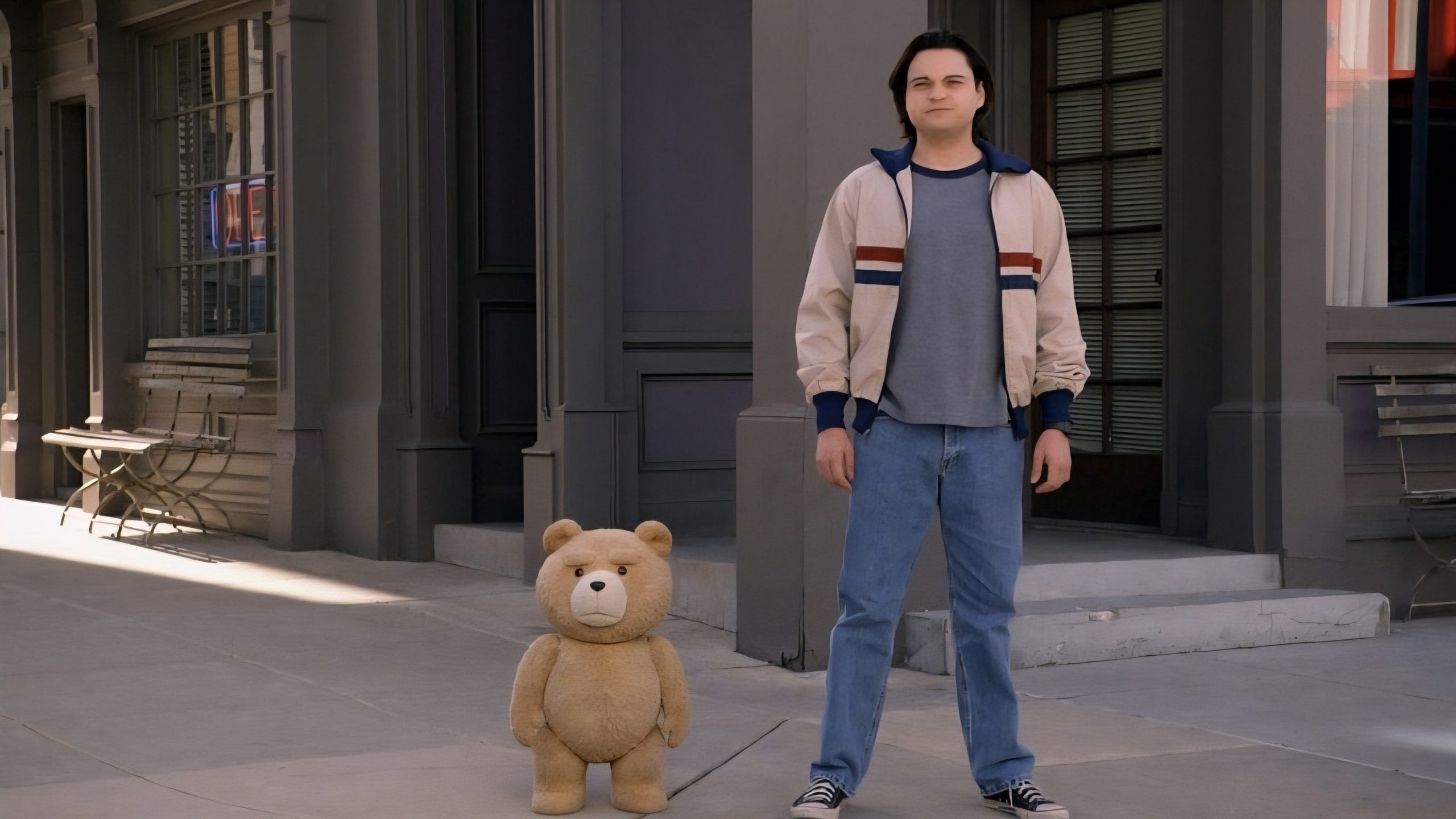 A young man in jeans and a jacket stands on a sidewalk next to a large, plush teddy bear in front of a building.