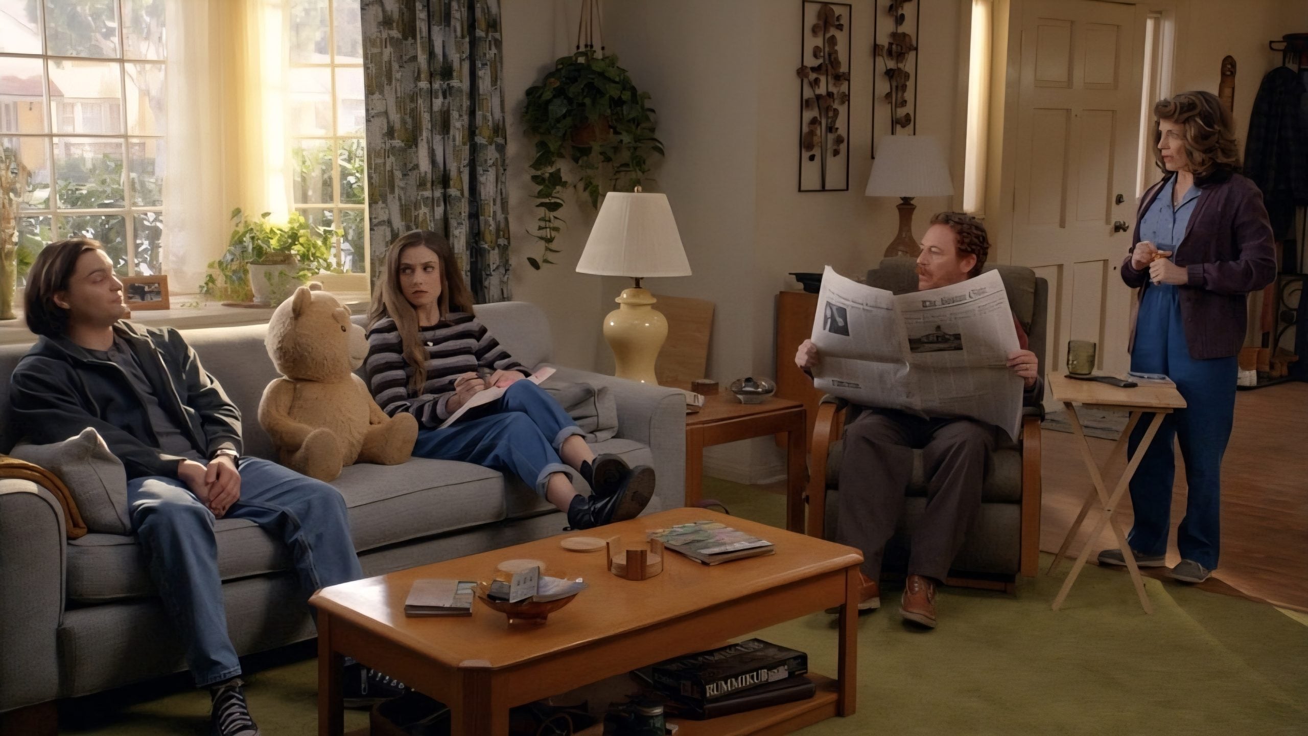 Four people sit in a living room; two are on a couch with a teddy bear, one reads a newspaper on a chair, and one stands holding a mug. The setting appears casual and domestic.
