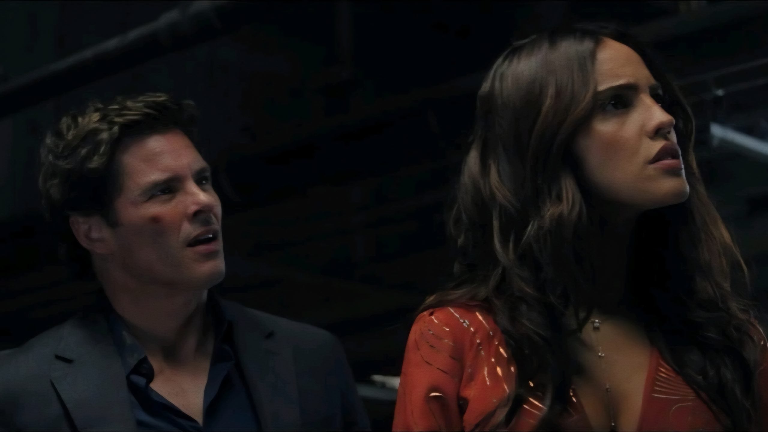 A man in a dark suit and a woman in a red patterned dress stand indoors, both looking toward something off camera with serious expressions.