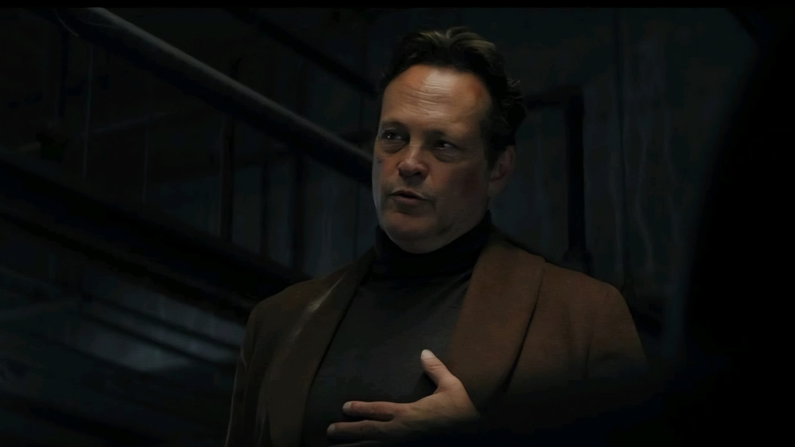 A man in a brown jacket and black turtleneck stands indoors with one hand on his chest, looking serious.