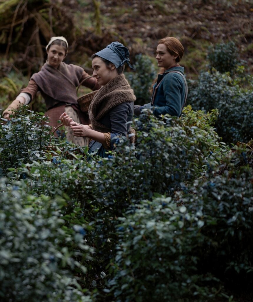 Three women wearing historical clothing pick leaves from dense green bushes in a forest setting.