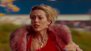 A woman with blonde hair, wearing a pink fur coat and colorful jewelry, appears to be running outdoors with a concerned expression.